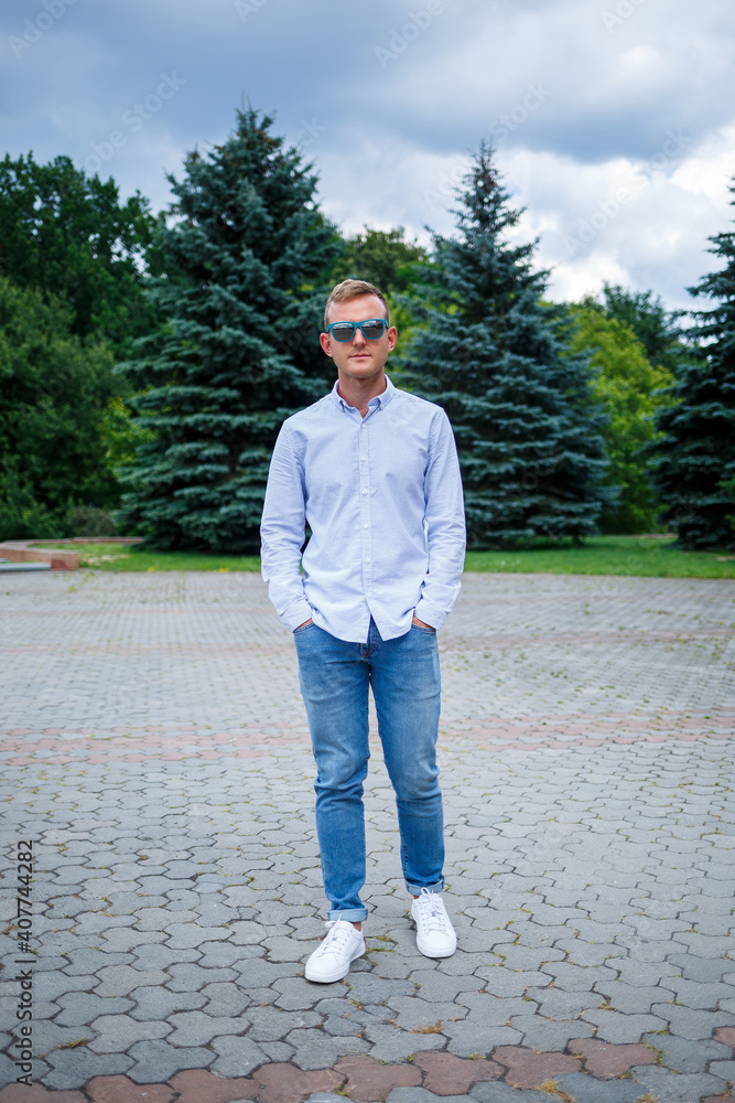 Fototapeta premium A handsome young man of European appearance wearing sunglasses is dressed in a shirt and jeans. The guy walks down the street, he is stylishly dressed