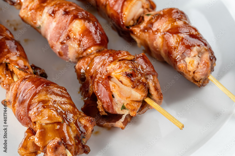 Grilled bacon-wrapped chicken tenders are laid out on a white plate. Strung on wooden skewers.