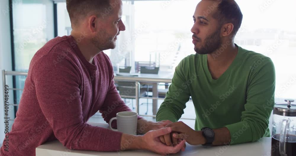 Multi ethnic gay male couple talking holding hands in kitchen Multi ethnic gay male couple talking holding hands in kitchen