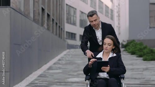 Crop view of man pushing wheelchair with his disabled colleague and communicating in businessarea. Female invalid person using tablet and telling direction to male person helping her. photo