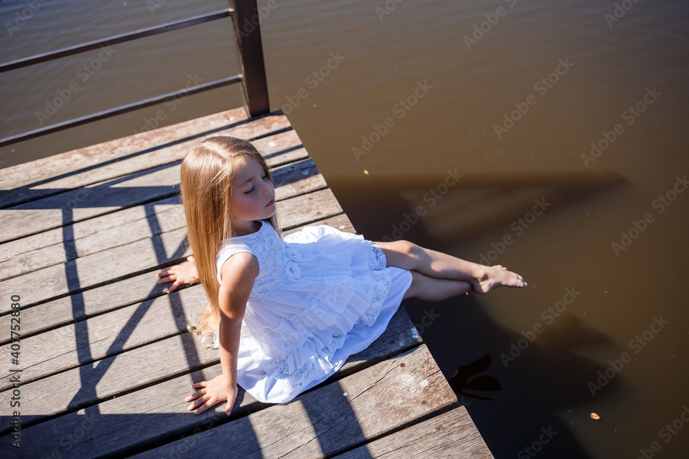 Fototapeta premium A 7-year-old girl with long blond hair by the lake sits on a clutch with legs in the water. She splashes her feet in the lake. Barefoot girl in a white dress with long hair.