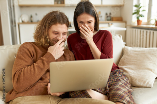 Portrait of cheerful young redheaded man sitting comfortably on sofa with his girlfriend laughing while watching stand up show together using generic laptop, enjoying weekend at home, having fun