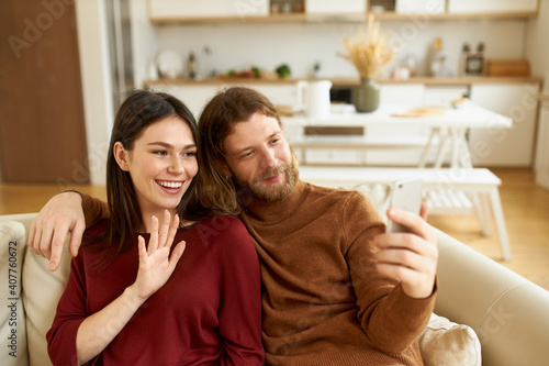 Handsome young bearded guy sitting in living room with his joyful wife, holding mobile phone, speaking to relatives on virtual chat, using online application, making greeting gesture with hand