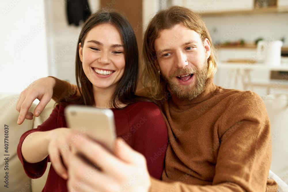 People, fun, leisure and modern technology. Cheerful relaxed young woman and bearded man entertaining themselves taking selfie using front facing camera on smart phone, making funny grimace People, fun, leisure and modern technology. Cheerful relaxed young woman and bearded man entertaining themselves taking selfie using front facing camera on smart phone, making funny grimace