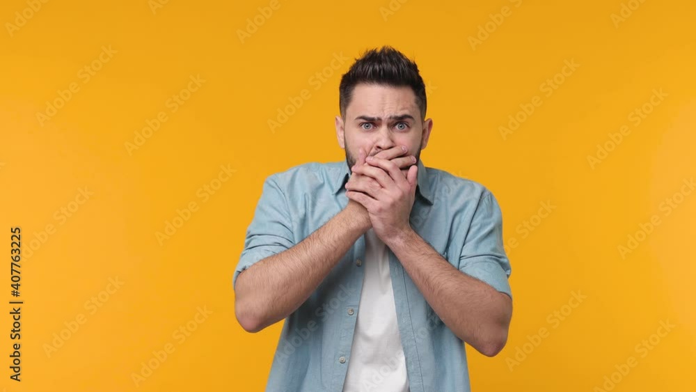 Shocked worried scared bearded young man 20s in casual blue shirt white t-shirt isolated on yellow color wall background in studio. People sincere emotions lifestyle concept. Covering mouth with hands Shocked worried scared bearded young man 20s in casual blue shirt white t-shirt isolated on yellow color wall background in studio. People sincere emotions lifestyle concept. Covering mouth with hands