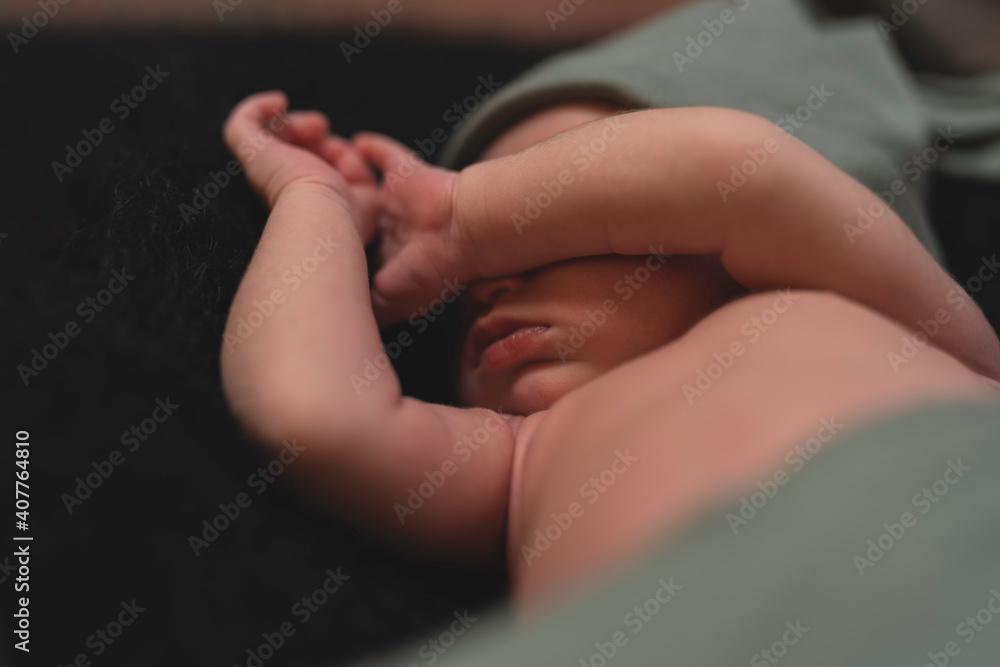 nfocused newborn 2 week old baby in a hat and covered with an olive-colored diaper covers his face with his hand on a black background