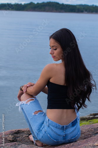 Vertical shot of a beautiful Hispanic female with long hair sitting on the coast rocks photo