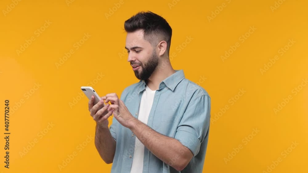 Amazed bearded young man 20s years old in casual blue shirt isolated on yellow background studio. People lifestyle concept. Pointing index finger on mobile phone browsing showing thumb up like gesture Amazed bearded young man 20s years old in casual blue shirt isolated on yellow background studio. People lifestyle concept. Pointing index finger on mobile phone browsing showing thumb up like gesture