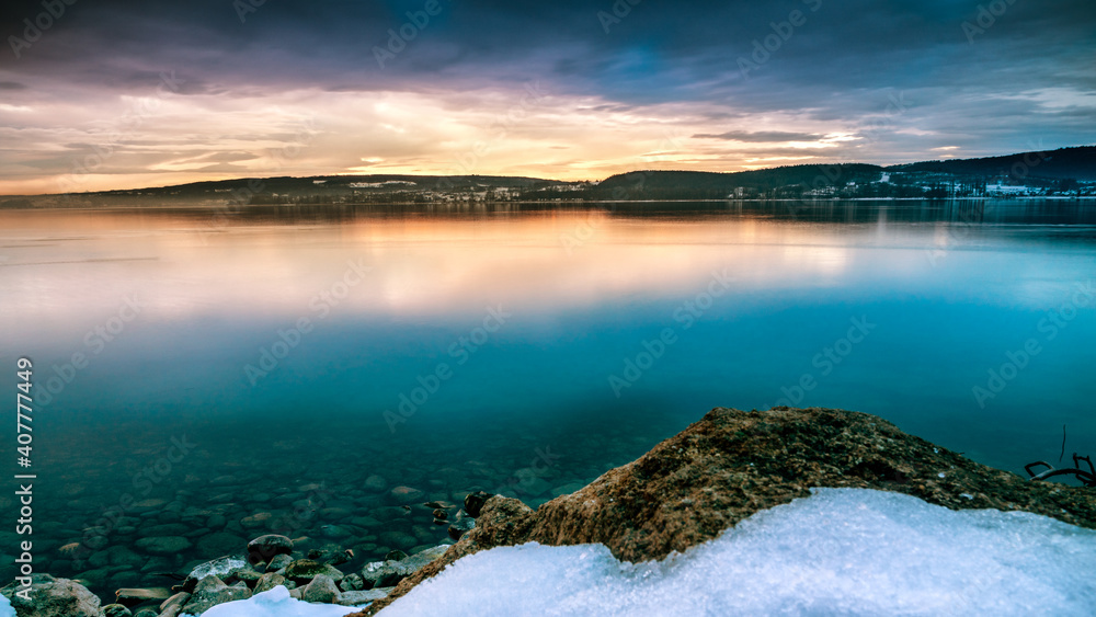 Wunderschöner Sonnenaufgang am Bodensee mit Schnee am Seeufer und Steinen am Wasser 