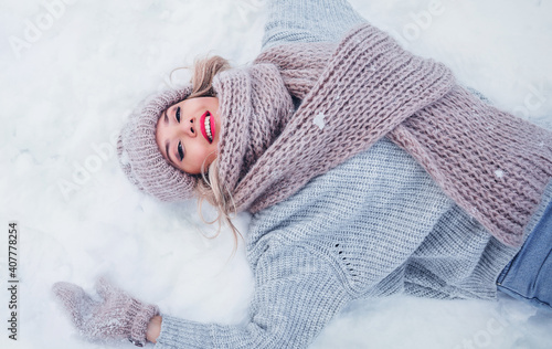 Winter fashion lifestyle portrait of beautiful young woman lying on the snow. Smiling and having fun. Wearing oversized sweater, scarf and mittens. Winter fun. Love and happiness concept. 