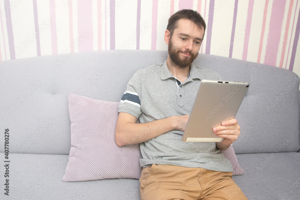 Attractive young man is using a tablet and smiling on sofa at home Attractive young man is using a tablet and smiling on sofa at home
