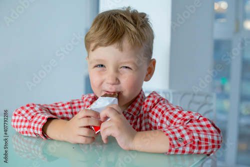 Boy sitting in the kitchen with a chocolate bar in his hands. Little boy eating delicious chocolate in the kitchen. 