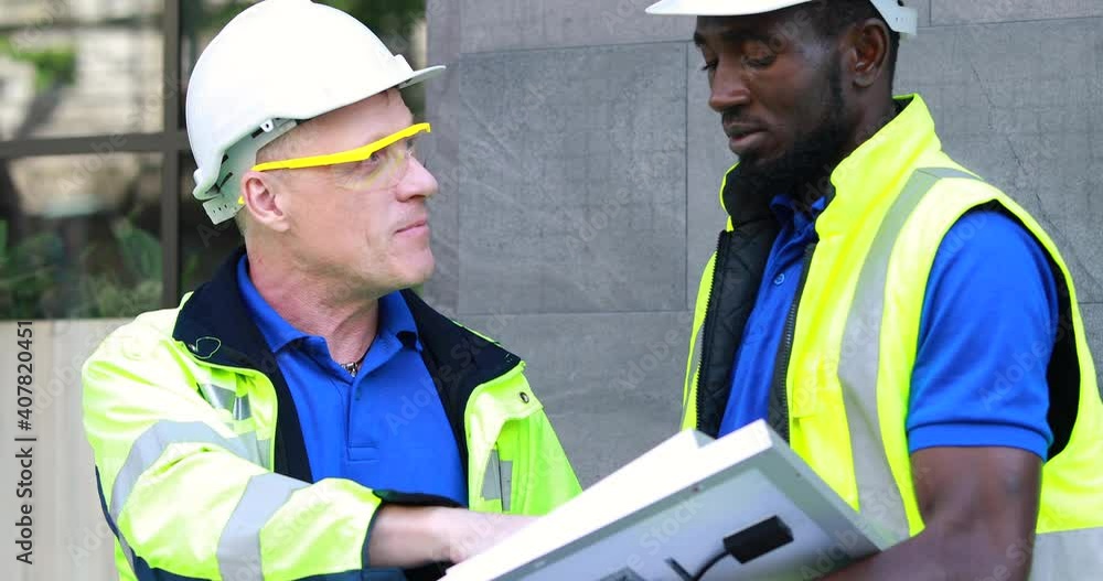 Two engineers African american engineer and caucasian electrician wearing white hard hat walk in new building holding solar panel on hand and Discuss Work Two engineers African american engineer and caucasian electrician wearing white hard hat walk in new building holding solar panel on hand and Discuss Work