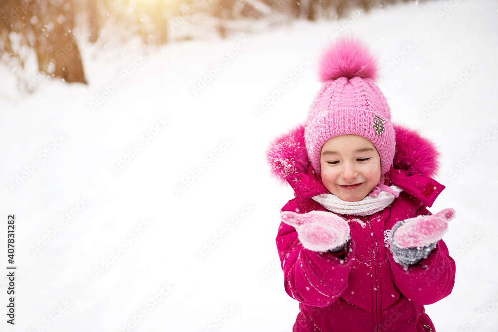Little girl in the cold looks at the snow mittens and smiles. Winter, walk the baby in the open air, snow. Warm clothing, knitted hat, fur pompom and hood. Snowball fight, winter fun, spring is coming Little girl in the cold looks at the snow mittens and smiles. Winter, walk the baby in the open air, snow. Warm clothing, knitted hat, fur pompom and hood. Snowball fight, winter fun, spring is coming