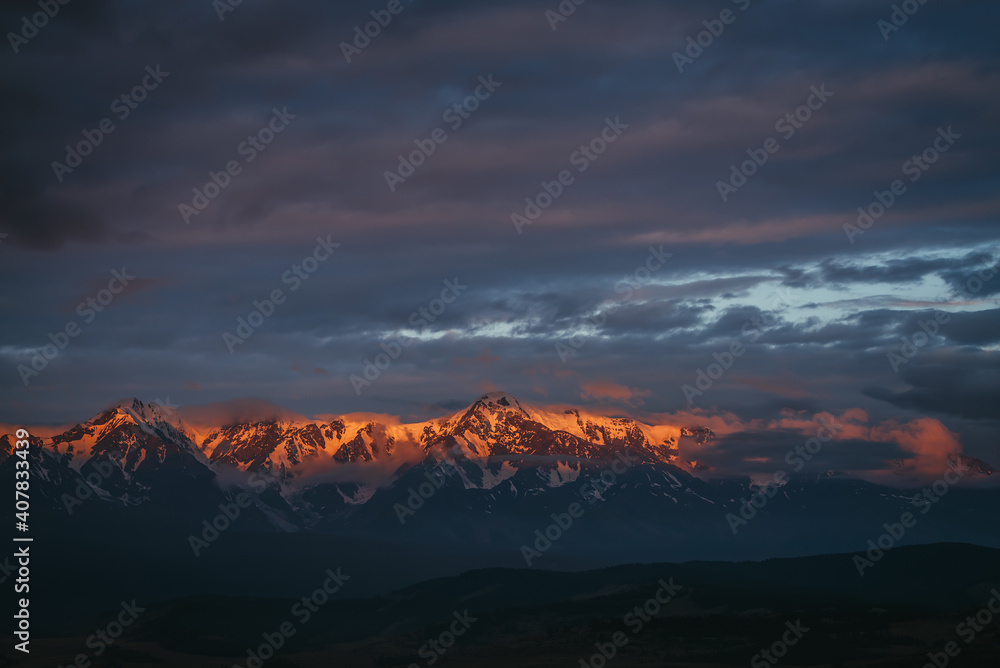 Scenic mountain landscape with great snowy mountain range lit by dawn sun among low clouds. Awesome alpine scenery with high mountain ridge at sunset or at sunrise. Big glacier on top in orange light.
