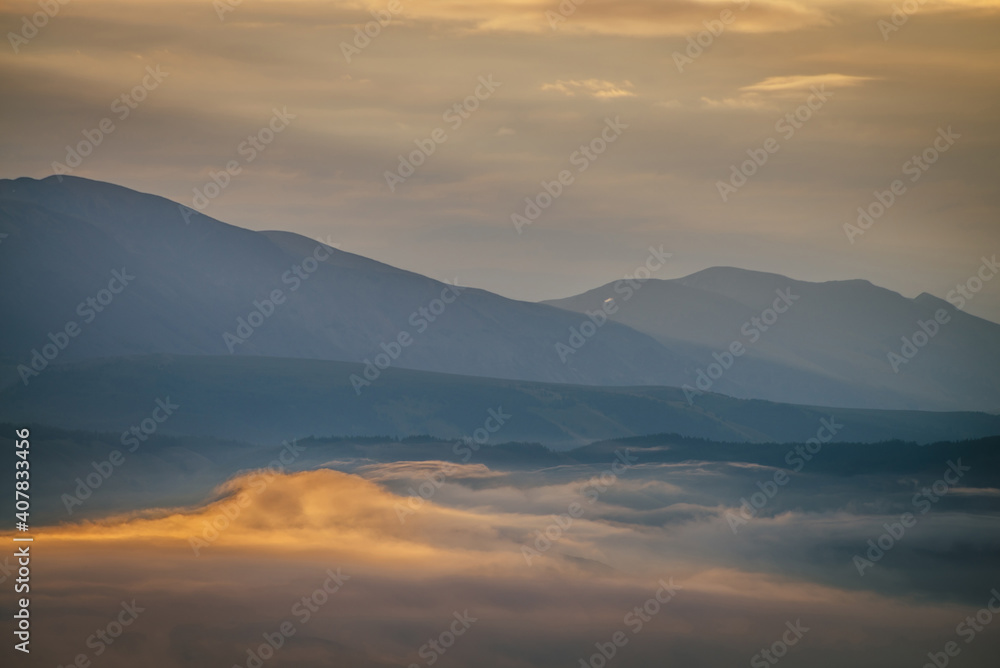 Scenic dawn mountain landscape with golden low clouds in valley among blue mountains silhouettes under cloudy sky. Sunset or sunrise scenery with low clouds in mountain valley in illuminating color.