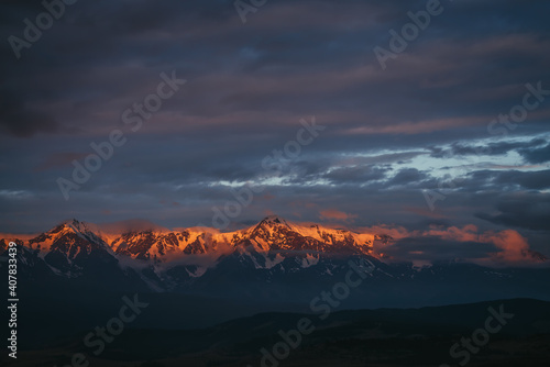 Scenic mountain landscape with great snowy mountain range lit by dawn sun among low clouds. Awesome alpine scenery with high mountain ridge at sunset or at sunrise. Big glacier on top in orange light.