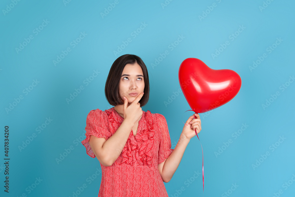 a doubting, hesitating young Asian woman in a red dress looks sideways, holds a flying red heart-shaped balloon and rubs her chin with her hand , isolated on a blue background.