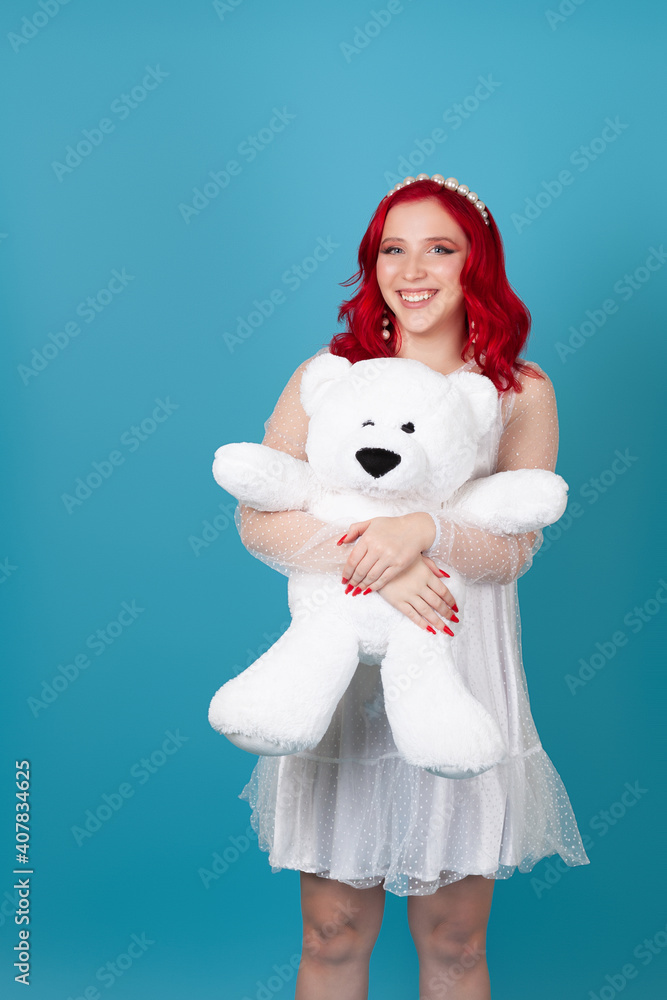 happy cheerful young woman woman with red wavy hair in white dress hugs big white teddy bear isolated on blue background. happy cheerful young woman woman with red wavy hair in white dress hugs big white teddy bear isolated on blue background.