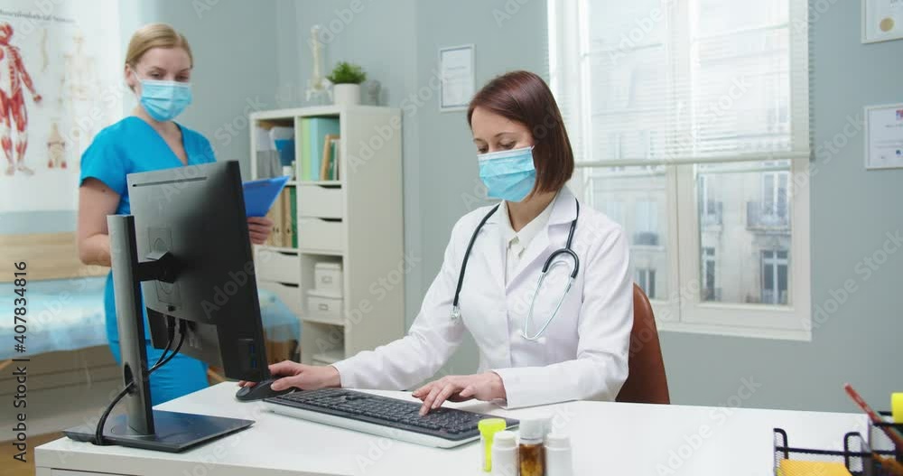 Adult female doctor in protective mask working on computer in hospital office. Pretty young nurse in blue uniform entering room and asking boss to check medical reports. Medicine, pandemic, health. Adult female doctor in protective mask working on computer in hospital office. Pretty young nurse in blue uniform entering room and asking boss to check medical reports. Medicine, pandemic, health.