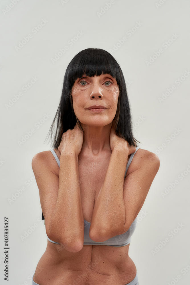Aging process. Vertical shot of attractive half naked middle aged caucasian woman looking at camera while posing in lingerie in studio, standing against grey background Aging process. Vertical shot of attractive half naked middle aged caucasian woman looking at camera while posing in lingerie in studio, standing against grey background