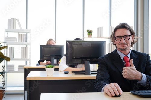 Portrait of businessman wearing suit thumb up and smiling with office at the background.