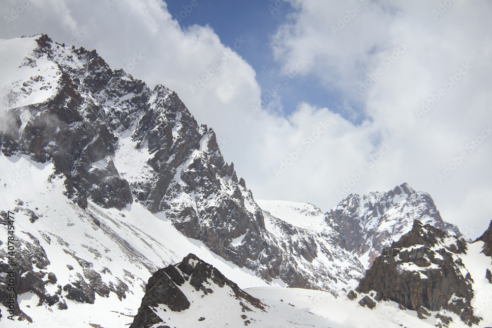 Fototapeta premium Alpine snow cliffs. Snowy cliffs high in the mountains, a clear winter sunny day, sharp peaks of the mountains in the snow against a blue sky with clouds, winter in the mountains