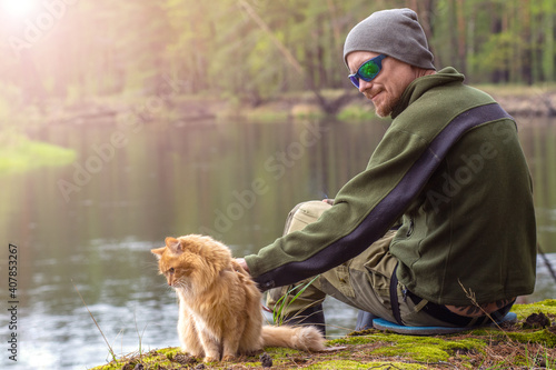 tourist man in a hat hugs a fluffy red cat by the river with sun glare