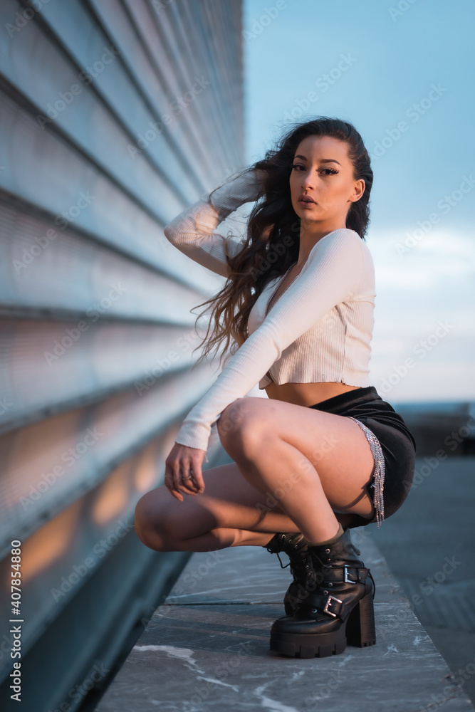 Posing of a brunette Arab girl crouching in the city at blue hour next to a gray building, wearing black boots and a black short skirt Posing of a brunette Arab girl crouching in the city at blue hour next to a gray building, wearing black boots and a black short skirt