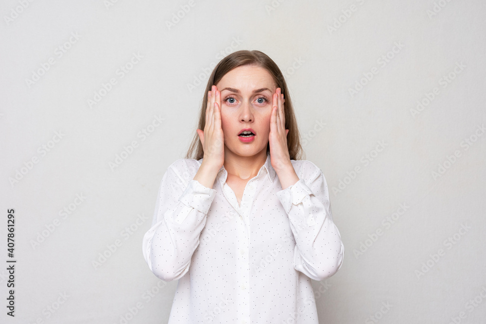 Portrait of a young woman clutching her head in fear, looking at the camera with her eyes wide open, white background Portrait of a young woman clutching her head in fear, looking at the camera with her eyes wide open, white background