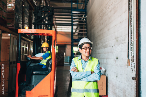 Portrait of asian man. warehouse Smiling worker standing with arms crossed in large warehouse with goods. in background driver at Warehouse forklift loader works with goods.