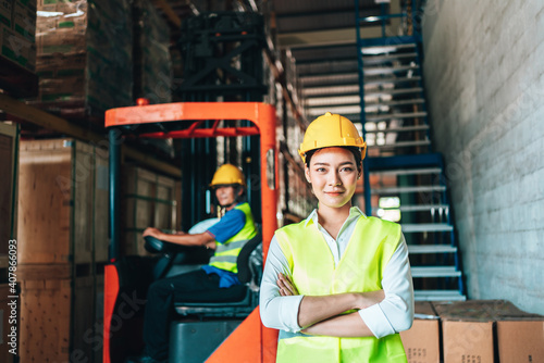 Portrait of asian woman. warehouse Smiling worker standing with arms crossed in large warehouse with goods. in background driver at Warehouse forklift loader works with goods.
