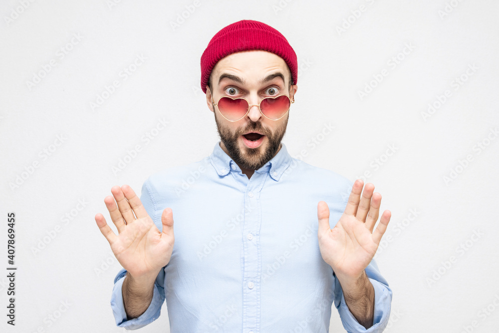 Dazed bearded lover shows stop sign with palms, man wears pink heart-shaped glasses, portrait, white background Dazed bearded lover shows stop sign with palms, man wears pink heart-shaped glasses, portrait, white background