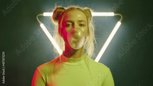 Camera zoom on modern young girl blowing gum bubble in changing colorful light on background with triangular neon lamp