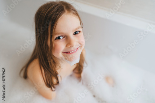 A small and beautiful red-haired girl, a child bathes, is washed in a white bath with soap foam. Happy childhood.