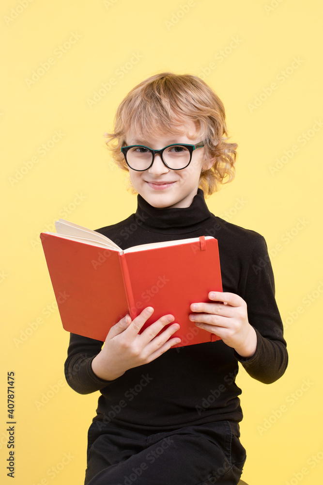 Blond schoolboy holding an open red book, smiling sweetly, wearing glasses, child in a black sweater. Copy space. Yellow background color trend 2021. Concept of poor eyesight, smart kid. Eye contact. Blond schoolboy holding an open red book, smiling sweetly, wearing glasses, child in a black sweater. Copy space. Yellow background color trend 2021. Concept of poor eyesight, smart kid. Eye contact.