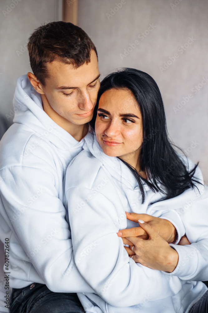 Beautiful young couple in the same white hoodie and black pants lie on the bed hugging. Dreaming of happy future together. Enjoying each other's company. Man gently hugs his beloved from back Beautiful young couple in the same white hoodie and black pants lie on the bed hugging. Dreaming of happy future together. Enjoying each other's company. Man gently hugs his beloved from back