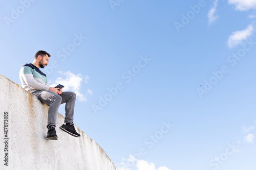 Funny Jump Free caucasian boy with a beard sitting on top of a wall with a tablet making a video call in sunlight outdoors and wearing colorful sweater 
