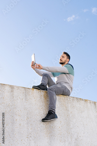 Funny Jump Free caucasian boy with a beard sitting on top of a wall with a tablet making a video call in sunlight outdoors and wearing colorful sweater 