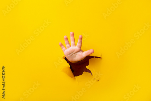 children's hand sticks out of a hole in a sheet of paper on a yellow background. Five finger sign, copy space