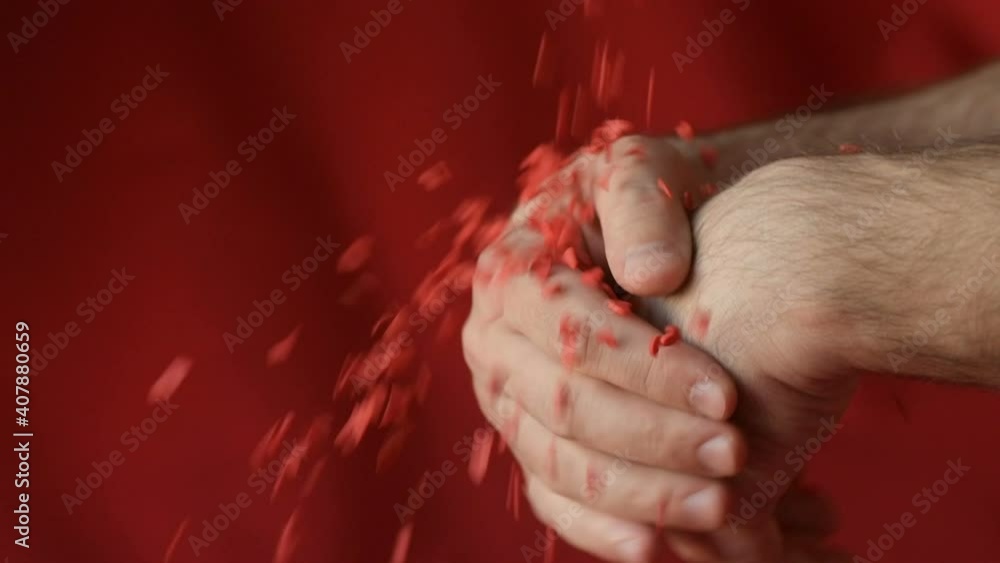 Red scarlet heart shaped sugar confetti crumbling and falling down in men's hands on red background. Romantic love, Saint Valentine's Day preparation design concept Red scarlet heart shaped sugar confetti crumbling and falling down in men's hands on red background. Romantic love, Saint Valentine's Day preparation design concept