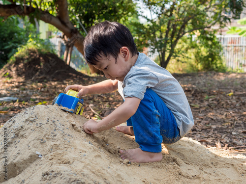 Asian child boy playing car toy on sand outdoor with cute face in nature background. Happy kid enjoy in relaxing day, preschool learning ef, family activity in summer, freedom concept.