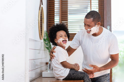 African American Man and little boy having fun laughing with shaving foam on their faces in bathroom at home. Funny Happy Black Father and son shaving faces in the morning