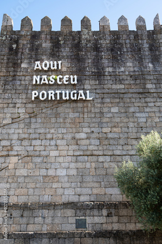 Medieval wall with the words “Here Portugal has born”. Tree in the foreground. Blue sky.