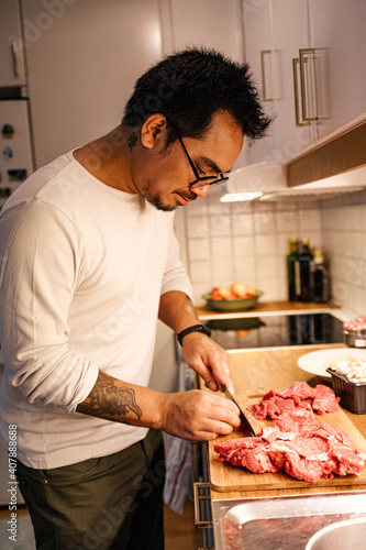 Young man cutting meat with a knife on a cutting board at home in the kitchen alone