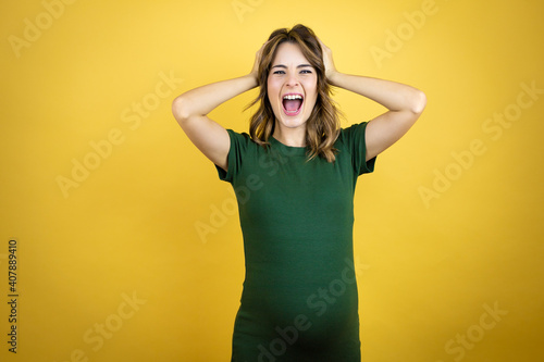 Young beautiful brunette woman pregnant expecting baby over isolated yellow background crazy and scared with hands on head, afraid and surprised of shock