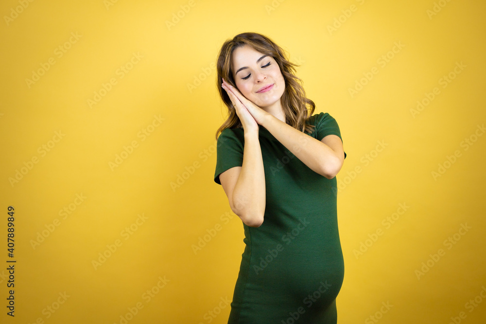 Fototapeta premium Young beautiful brunette woman pregnant expecting baby over isolated yellow background sleeping tired dreaming and posing with hands together while smiling with closed eyes.