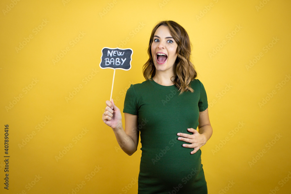 Young beautiful brunette woman pregnant expecting baby over isolated yellow background amazed and holding blackboard with new baby word message Young beautiful brunette woman pregnant expecting baby over isolated yellow background amazed and holding blackboard with new baby word message