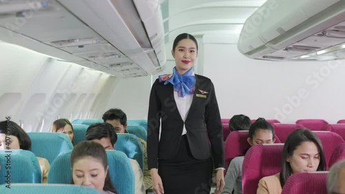 Portrait of attractive stewardess in uniform walking in the economy class of airplane and smile. Passengers sitting and fasten seat belt for safety flight. Transportation and service concept.