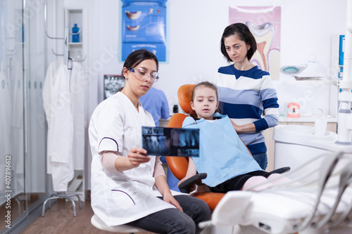 Little girl and parent listening dentist advice pointing at dental radiography. Stomatologist explaining teeth diagnosis to mother of child in health clinic holding x-ray.